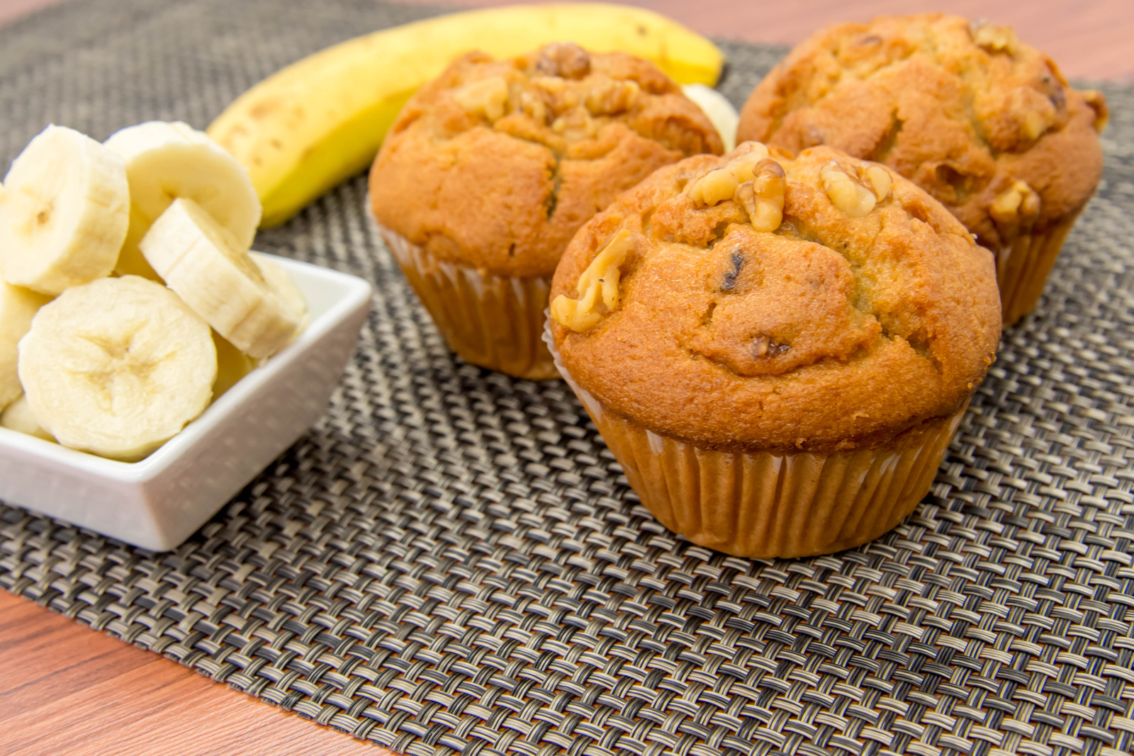 a whole banana lays in the back of a small square bowl of banana slices to the left of three perfectly baked banana nut muffins