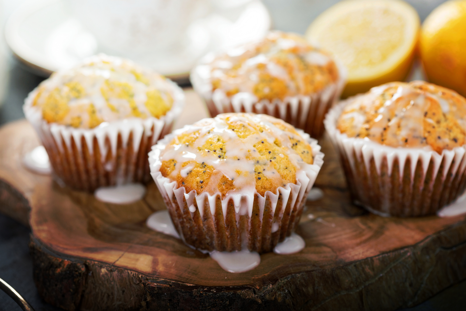 four lemon poppy seed muffins on a live-edge cutting board with icing dripping off them and some cut lemons in the background